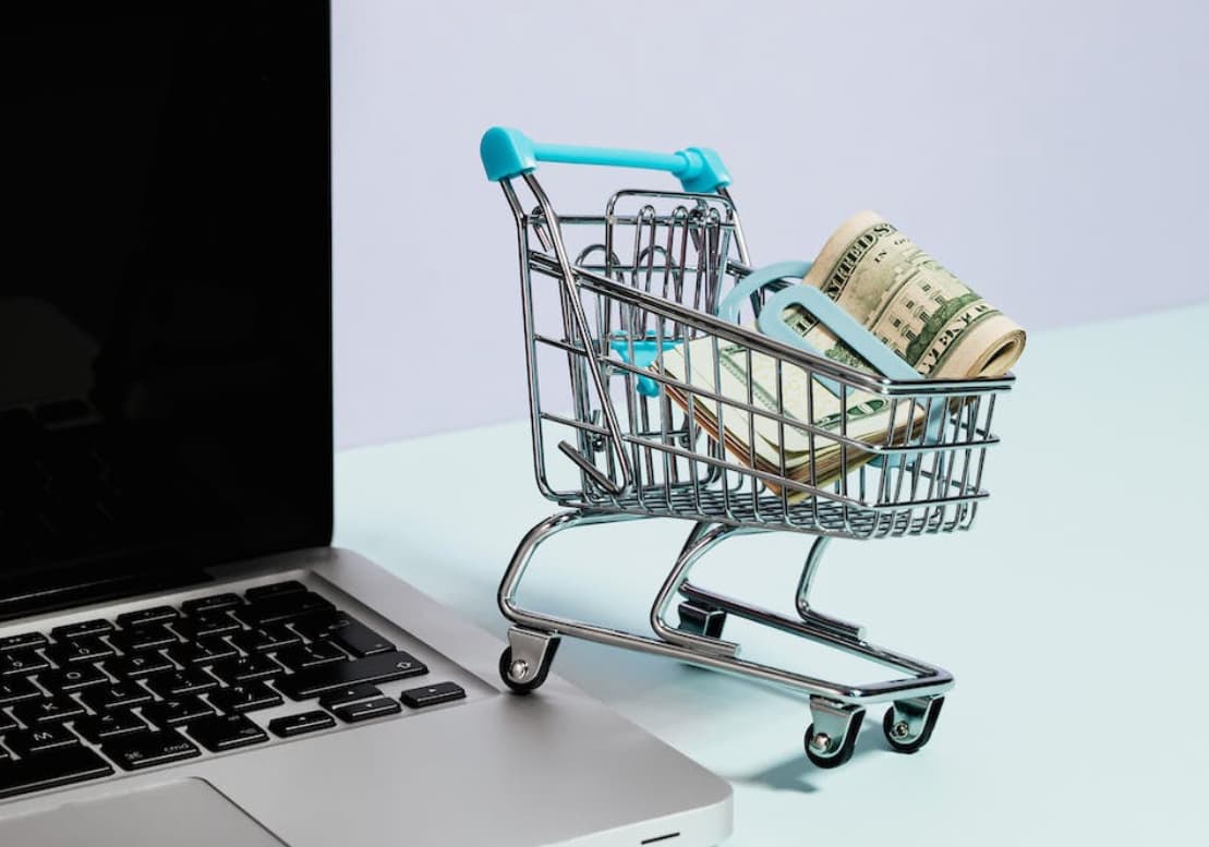 A shopping cart on a laptop keyboard filled with rolled-up banknotes, implying online shopping or digital finance